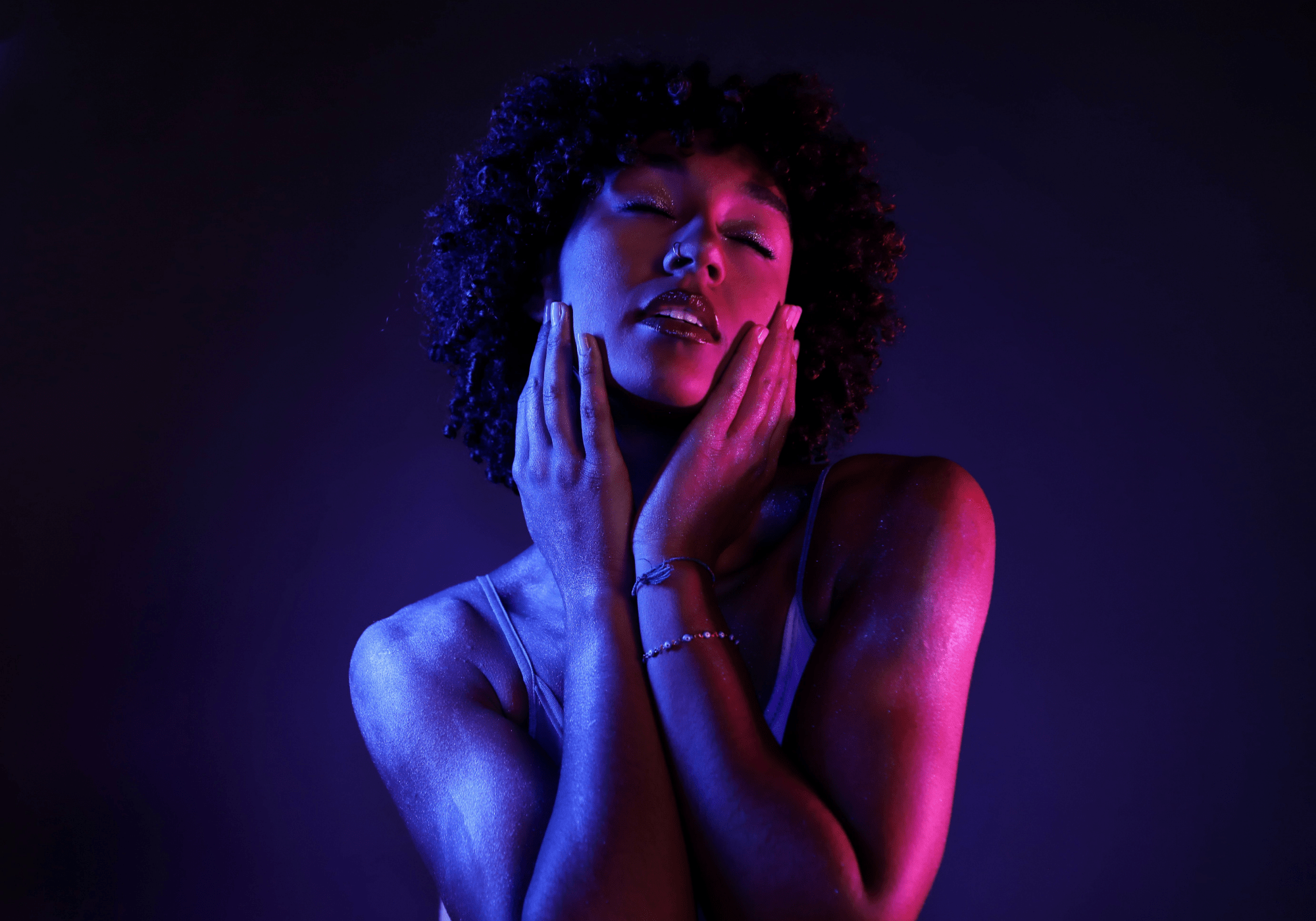 Close-up of a woman with curly hair, eyes closed, holding both hands to her cheeks under blue and pink lighting.